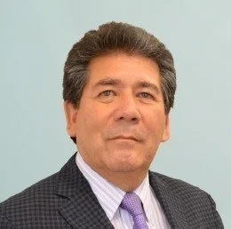 An older man with gray hair smiling, dressed in a dark pinstripe suit, blue shirt, red striped tie, and a matching pocket square.