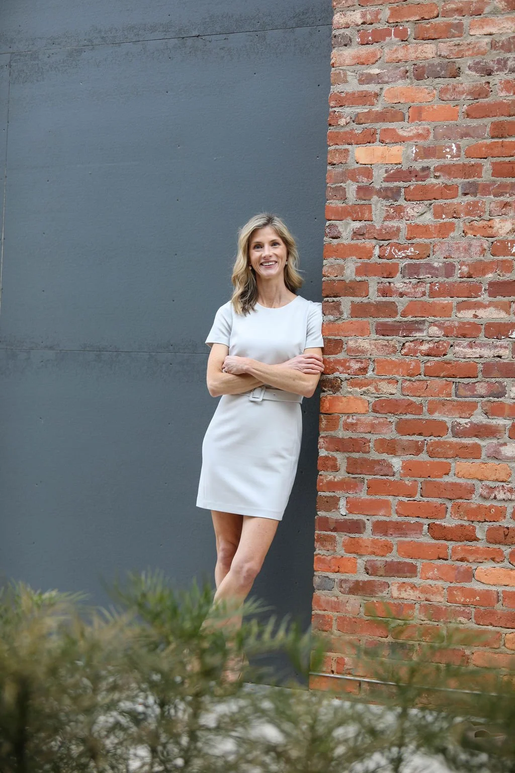 A woman in a white dress standing with arms crossed, smiling, beside a brick wall and a gray wall, with some greenery in the foreground.