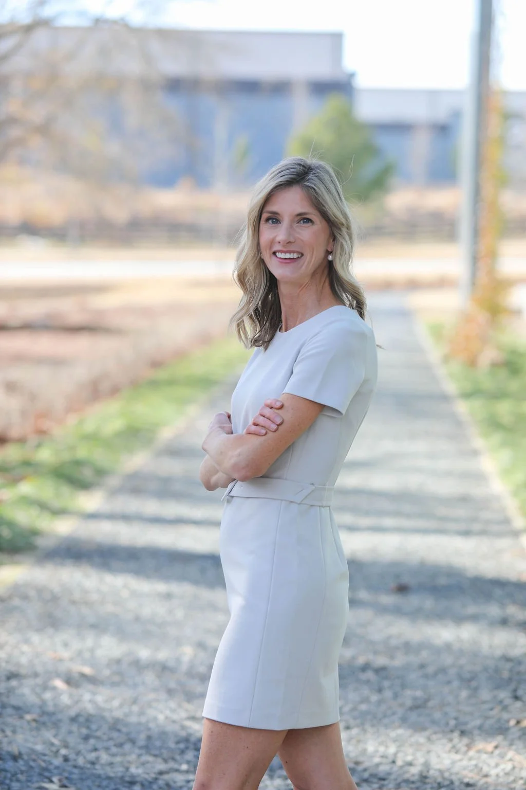 A woman in a white dress standing outdoors on a sidewalk with her arms crossed, smiling at the camera.
