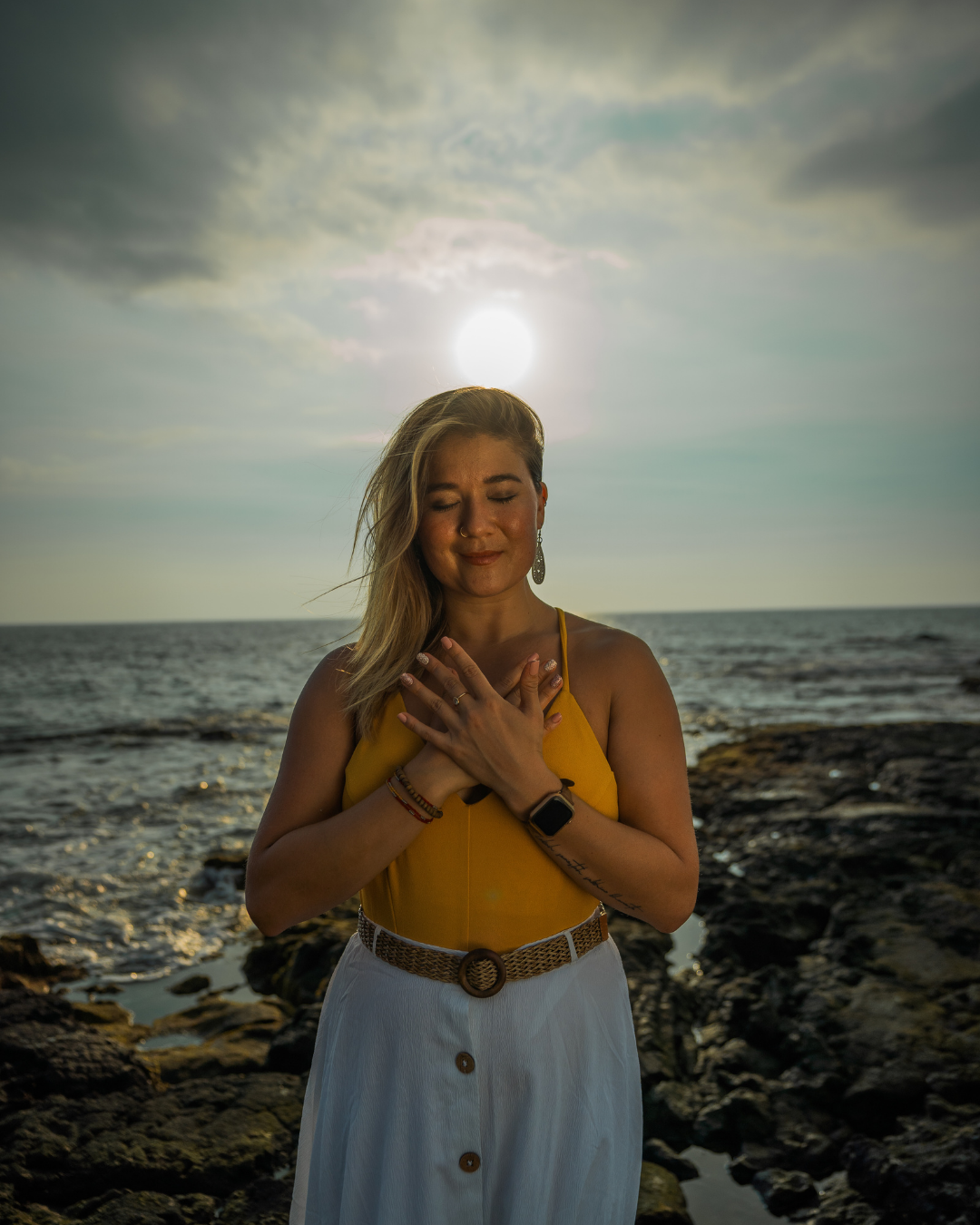A woman standing on rocky shore at sunset with hands crossed over her chest, eyes closed, smiling softly, wearing a yellow top and white skirt, with the sun and clouds in the sky behind her.