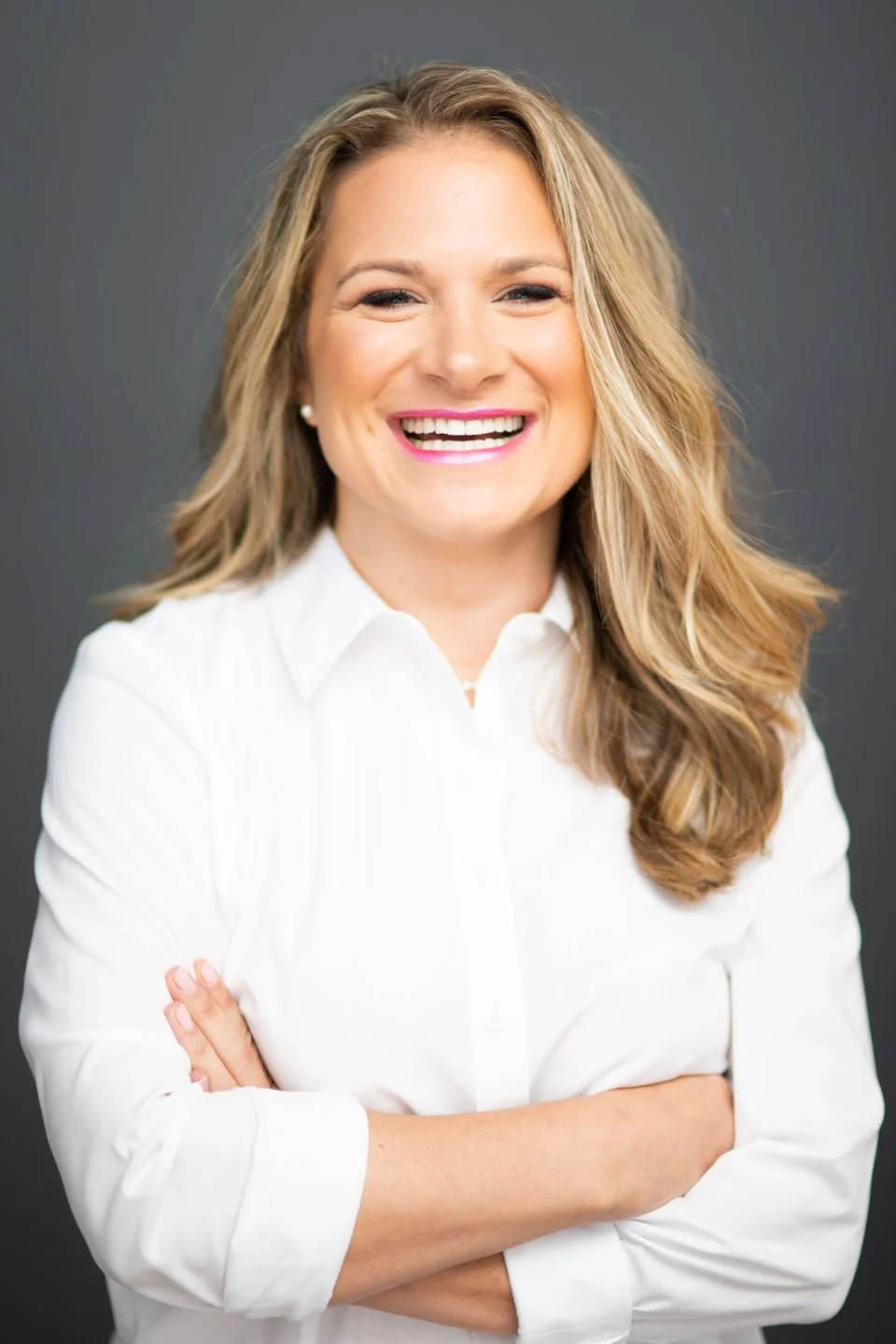 A woman with long, wavy blonde hair, smiling, wearing a white shirt with arms crossed, standing against a dark gray background.