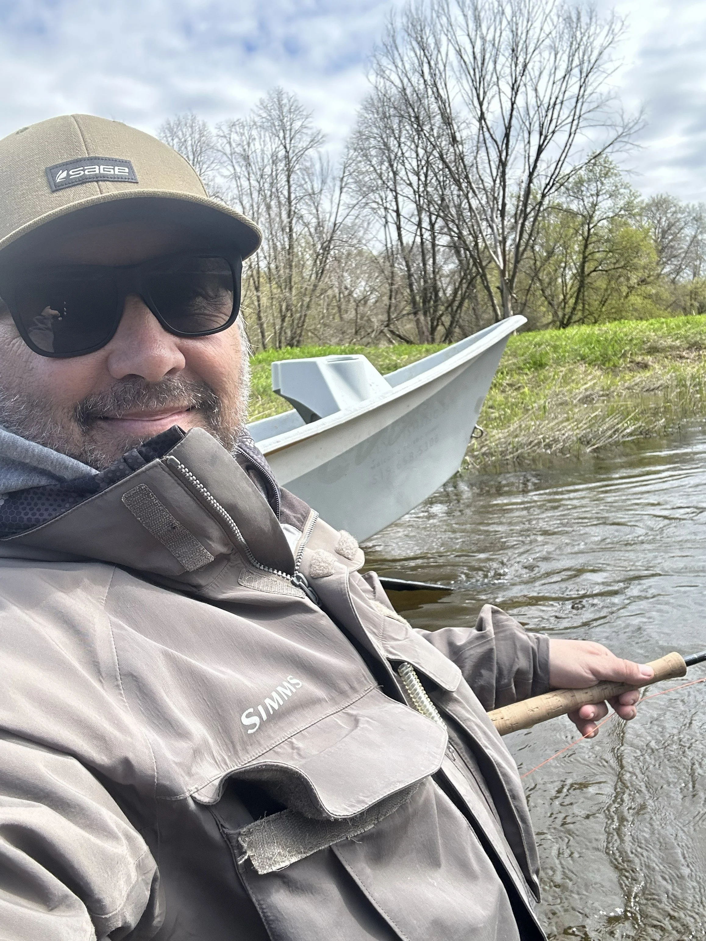 A man wearing a beige hat, sunglasses, and a gray jacket with the brand 'Sage' on it, holding a fishing rod near a body of water with trees in the background.