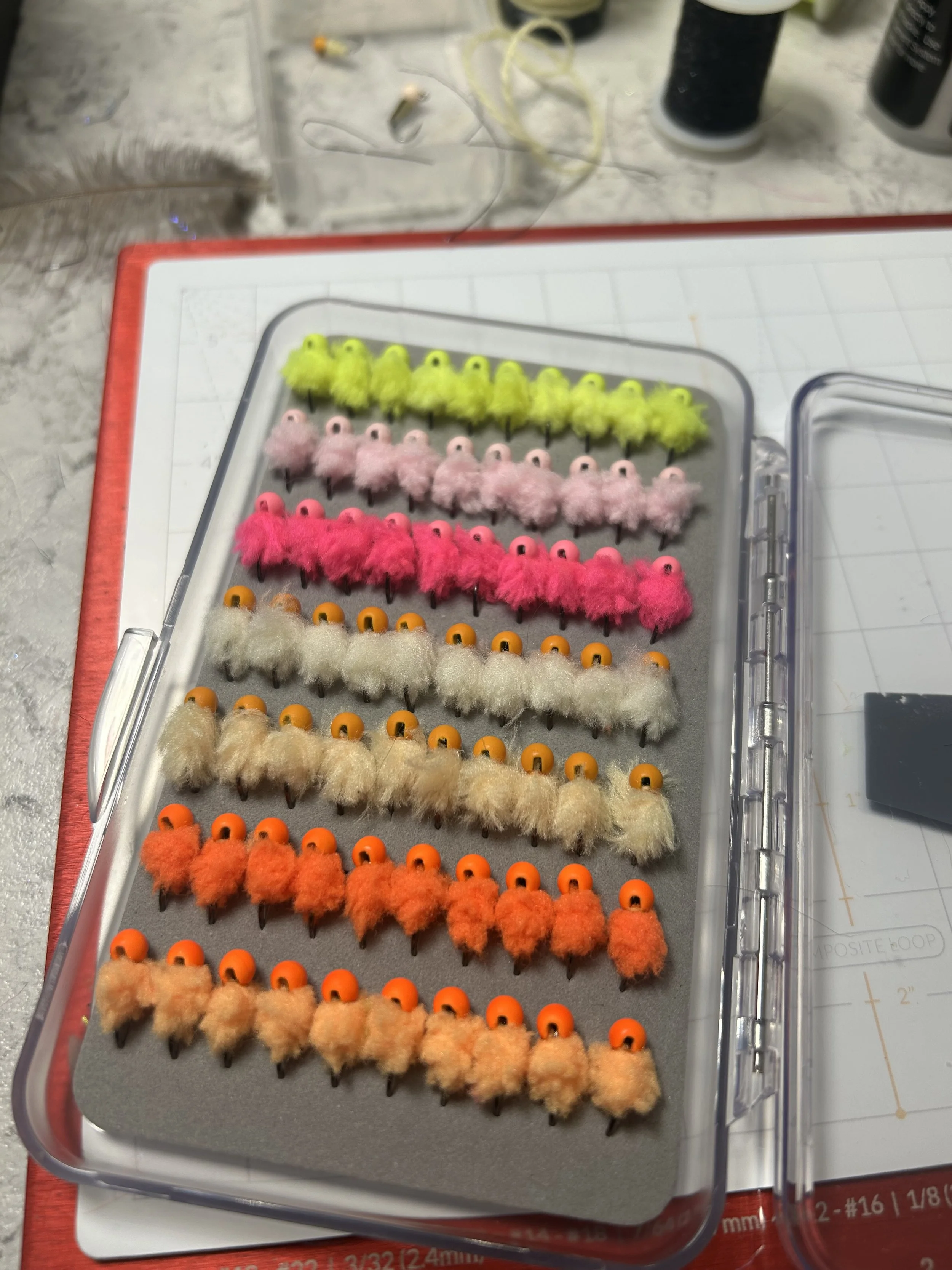 A plastic container with rows of colorful fuzzy fishing lures in shades of yellow, pink, white, orange, and beige, each with a small round hook at the bottom.