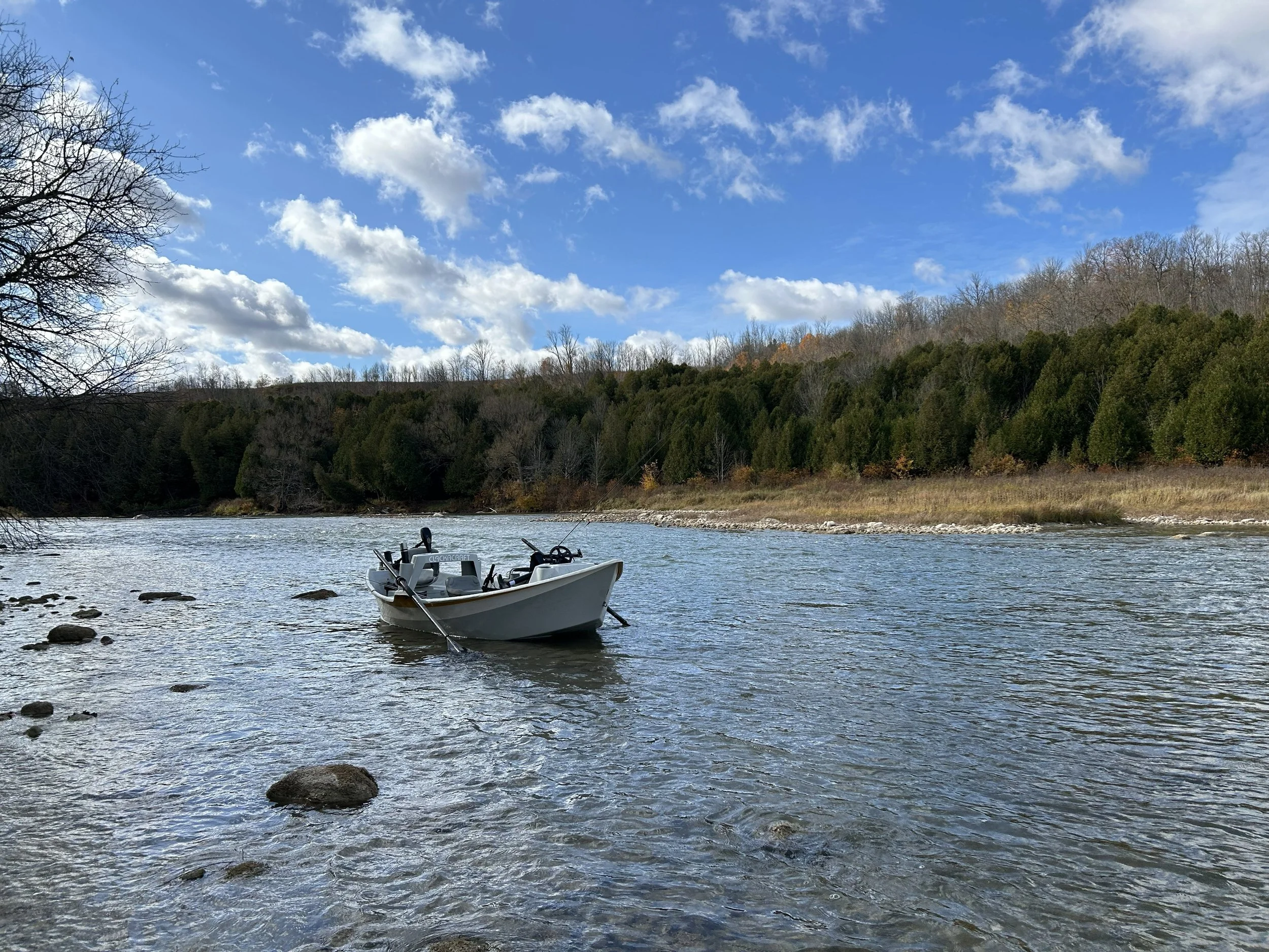 A small boat floating on a river with rocky shoreline, surrounded by leafless trees and dense green forest on a partly cloudy day.