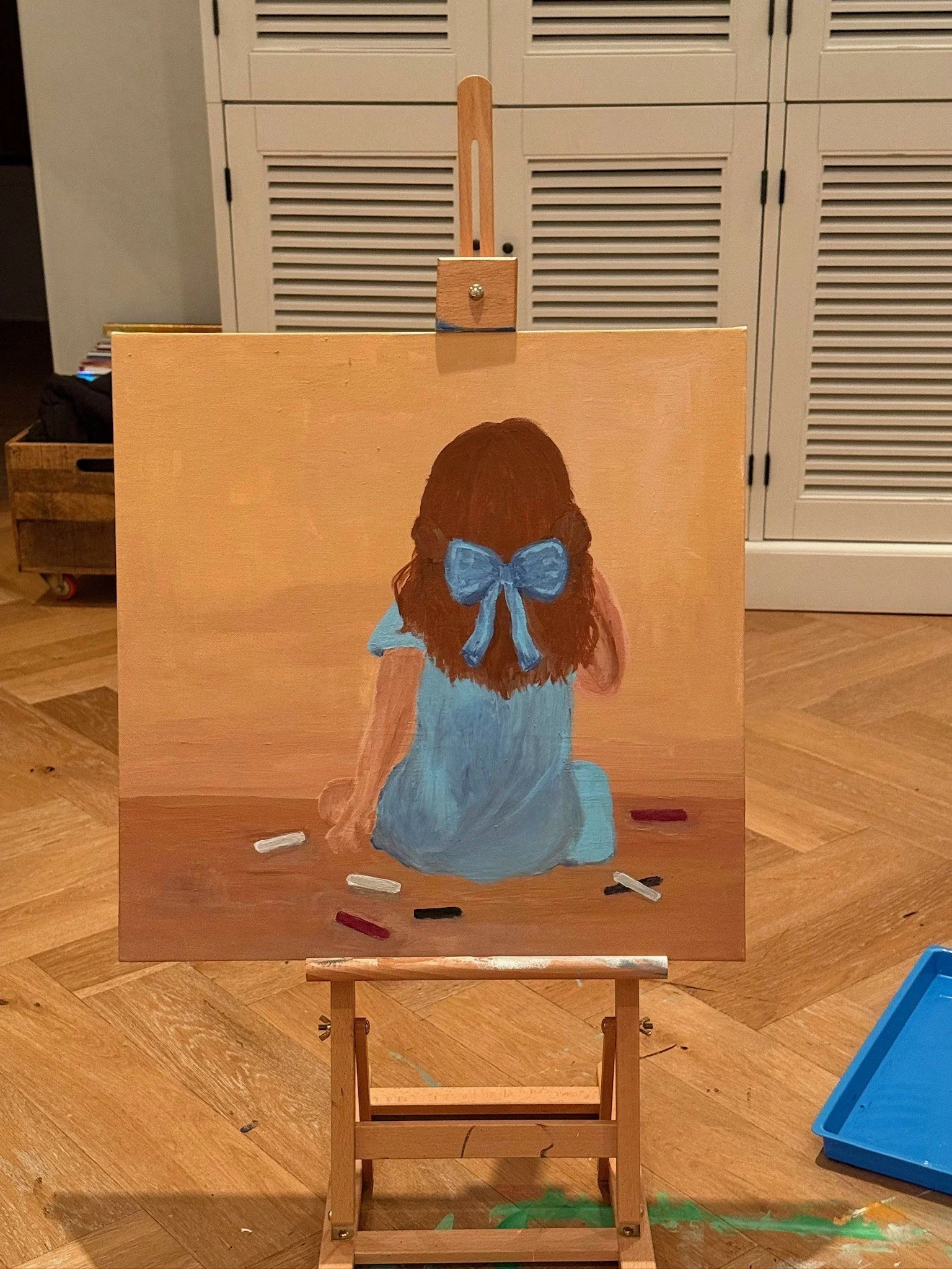 GIRLS WITH A BLUE BOW. Acrylic on canvas. 20×24". A child seen from behind, kneeling on a warm ochre floor with sticks of pastel scattered around her and a blue bow tied in her hair. Her back is turned, leaving both her face and the drawing she is ma