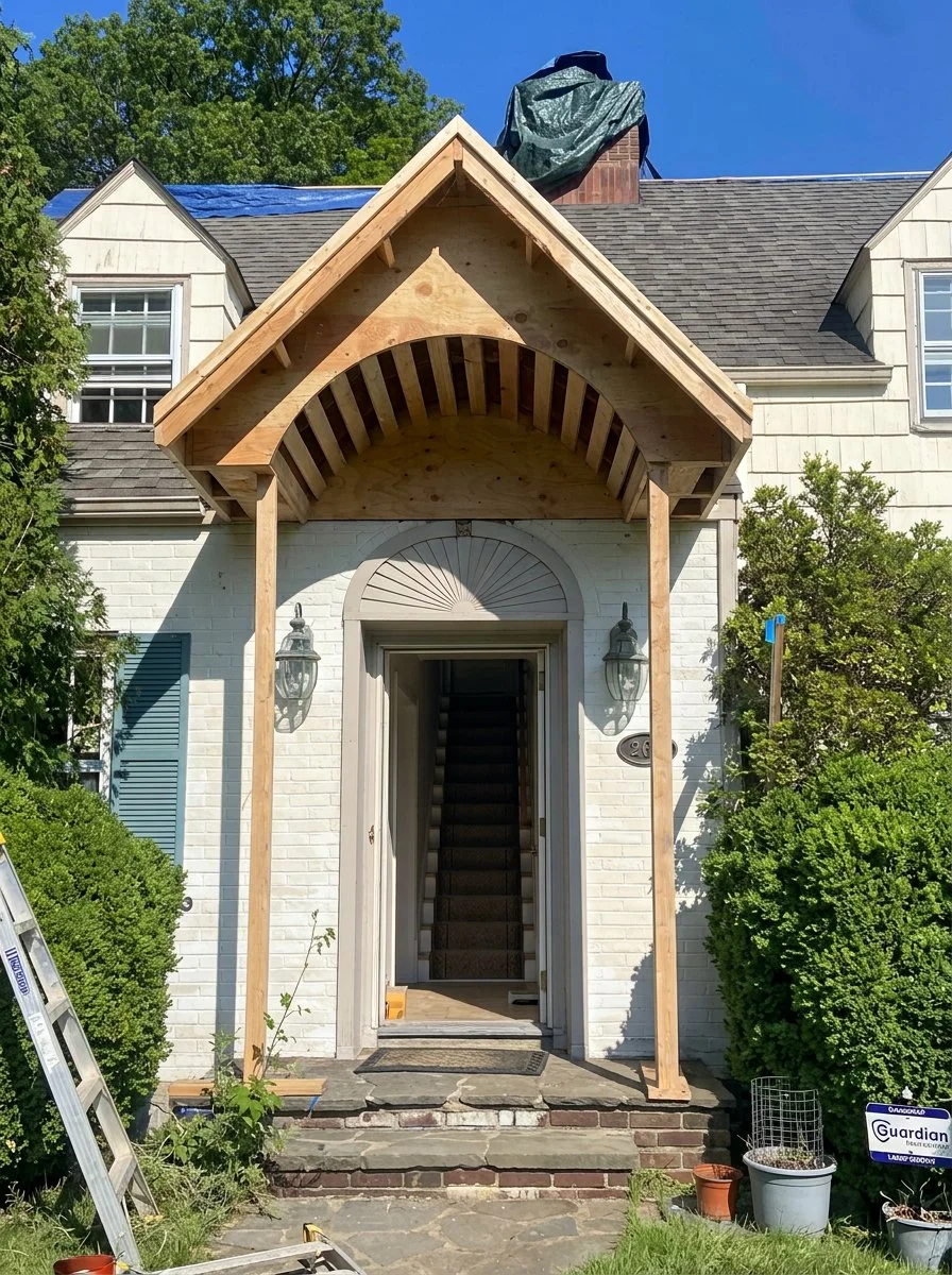 House entrance with a small wooden porch under construction, featuring a peaked roof, next to white brick walls and surrounded by green bushes, with a ladder and construction tools nearby.