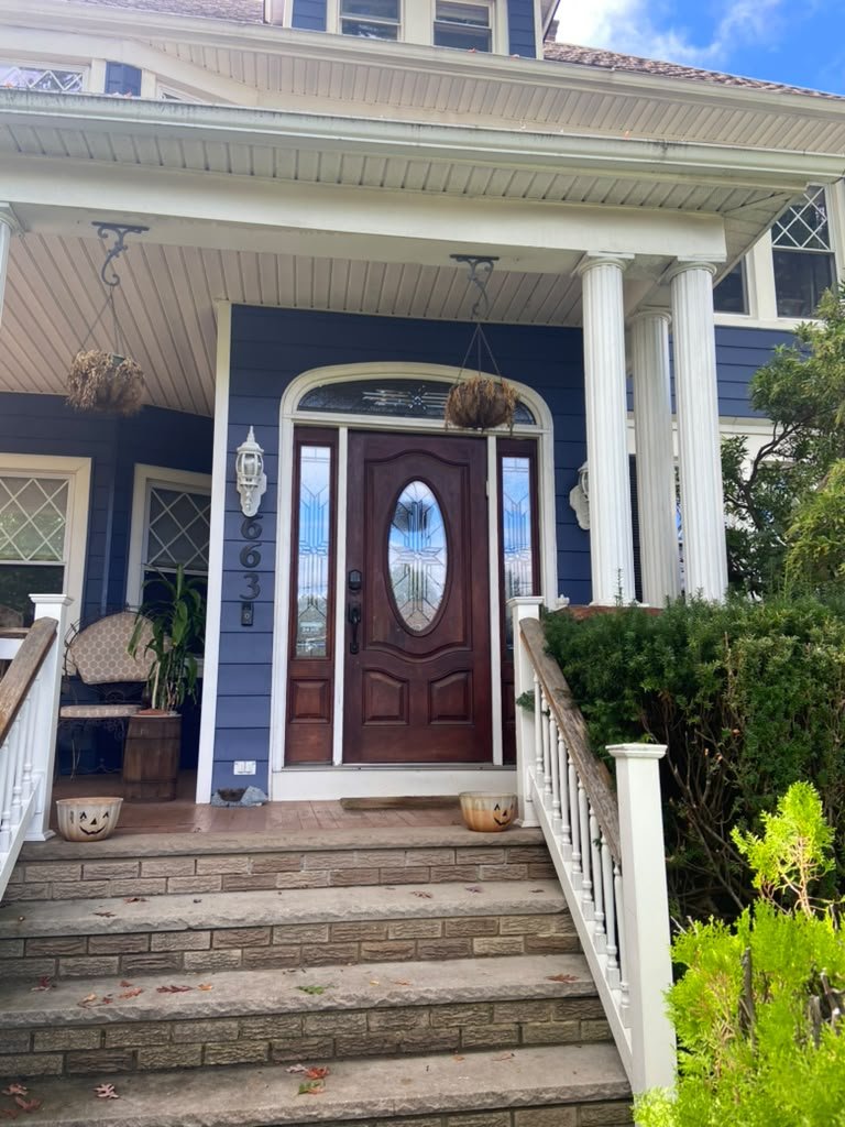 Front porch of a blue house with white columns and a brown wooden door with an oval glass window, decorative pumpkins on steps, and hanging plant baskets, with greenery in the front yard.