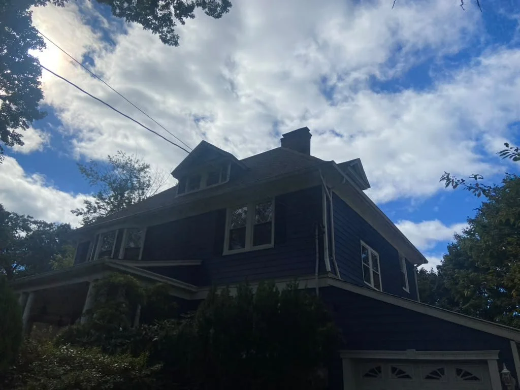 A two-story house with dark blue siding, white trim, and a steeply pitched roof with dormer windows, surrounded by trees and under a partly cloudy sky.