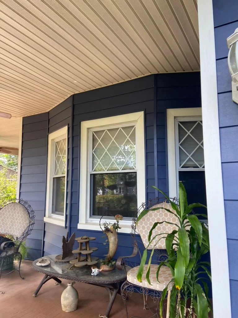Front porch of a house with blue siding, two decorative chairs, a glass table with ornaments, and a potted plant.