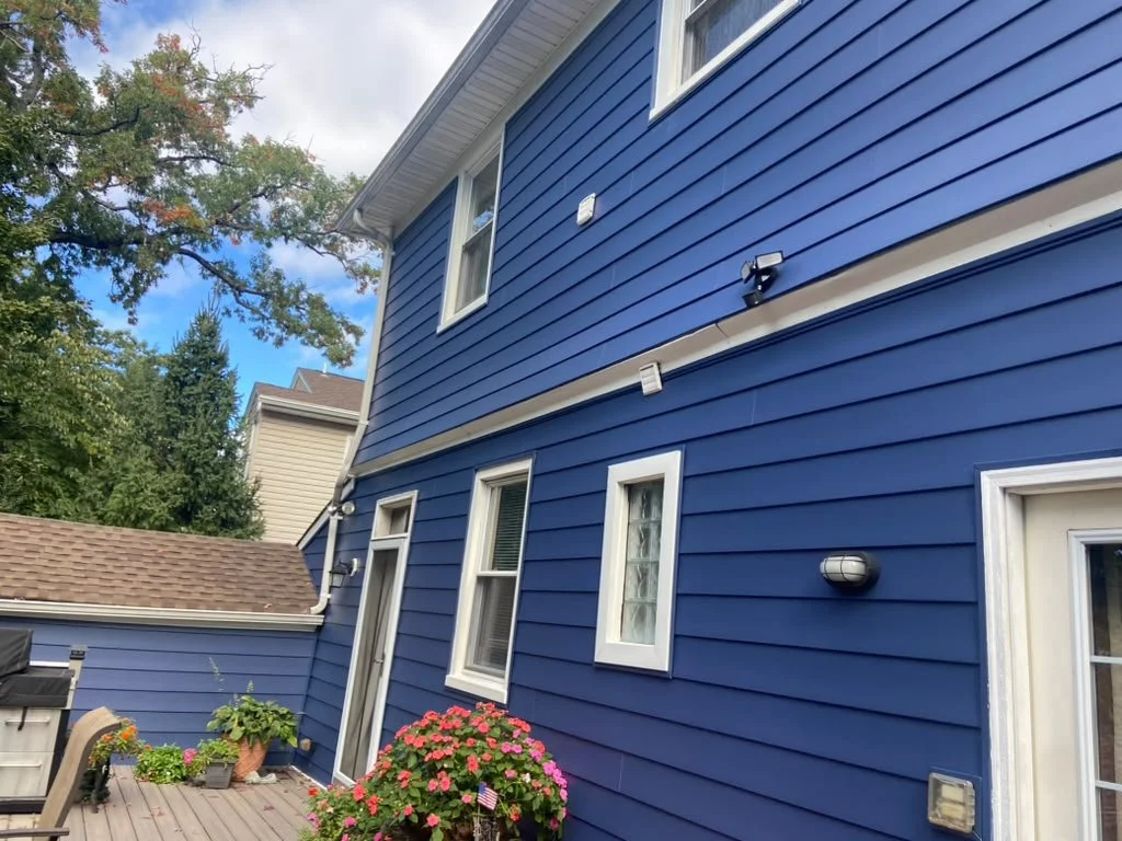 Backyard deck with blue house siding, white-framed windows, potted plants, and pink flowering bush, with neighboring houses and trees in the background.