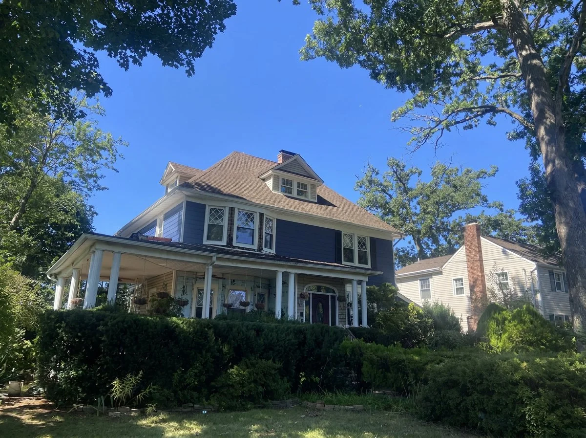 A large, two-story house with a brown roof and blue siding, surrounded by trees and bushes, under a clear blue sky.