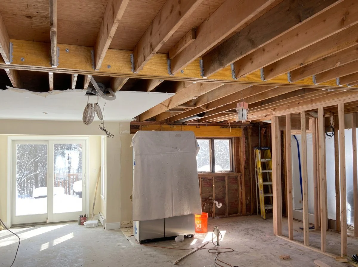 Interior of a home under renovation, with exposed wooden beams, construction materials, and equipment, including a ladder, a covered appliance, and a sliding glass door leading to snow-covered outdoors.