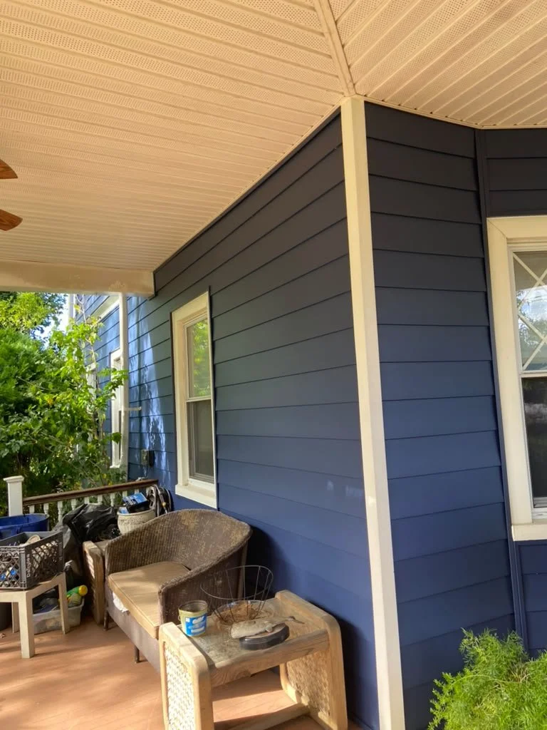 A porch with a blue house exterior wall, a window, a small wicker bench, a side table with a can, and a variety of items stored on shelves.