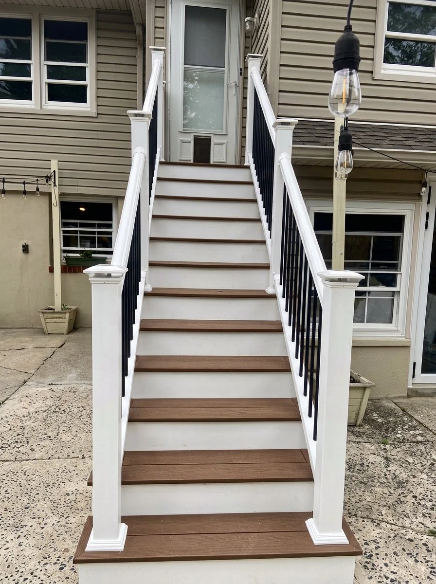An outdoor staircase with white risers, brown treads, black railings, leading up to a front door of a house with beige siding and windows.