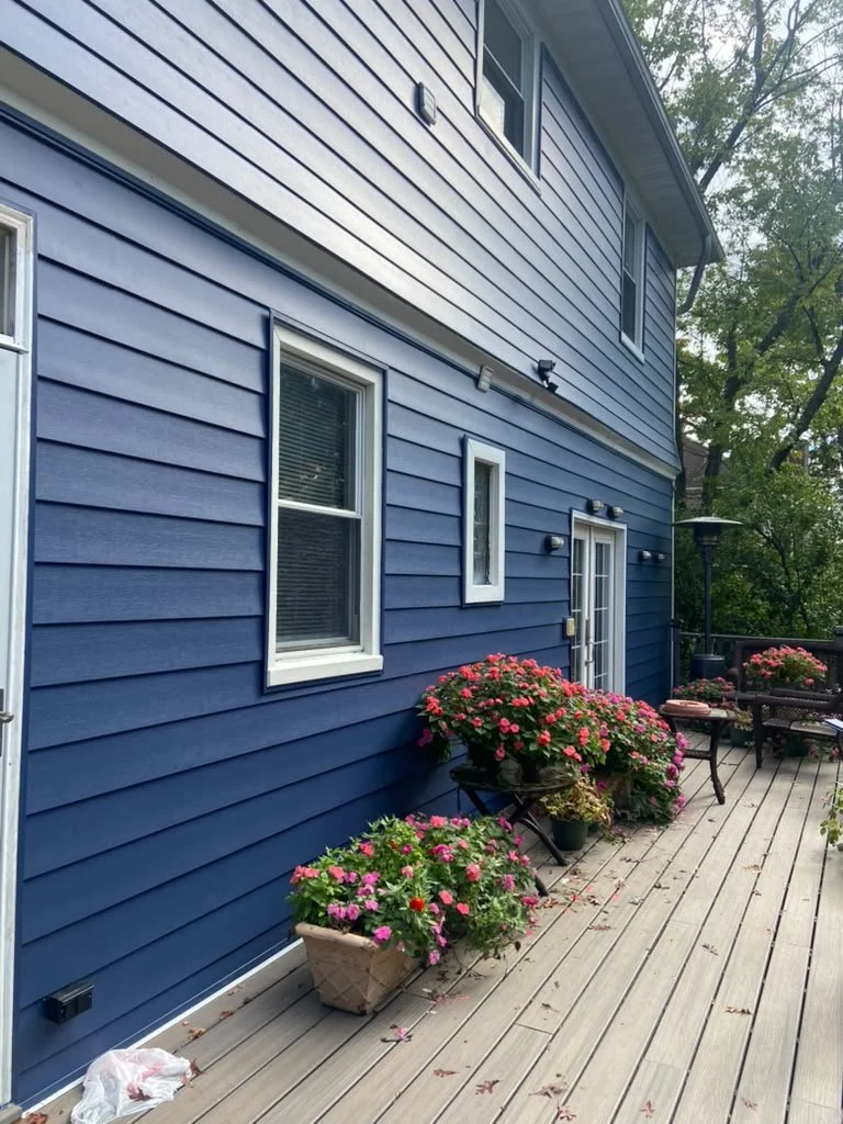 Blue house exterior with white-trimmed windows and door, decorated with pink and red flowers on a wooden porch.