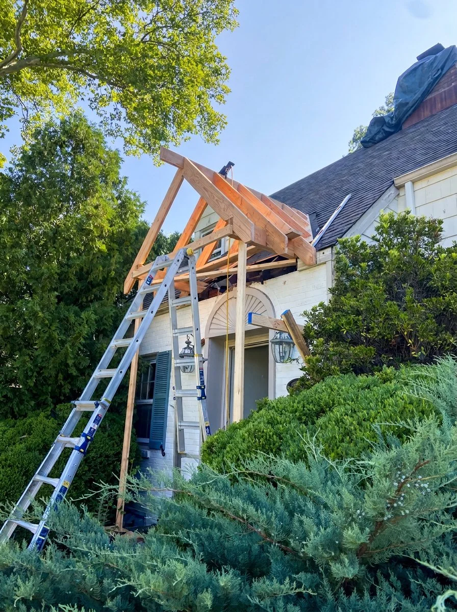 Construction project on the roof of a house, with a ladder leaning against the house and wooden framing in progress. Green bushes and trees surround the house on a sunny day.
