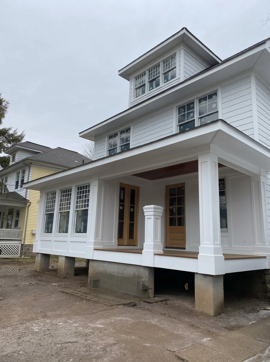A white, three-story house under construction with a front porch supported by columns, multiple windows, and wooden doors, on a dirt lot with neighboring houses in the background.