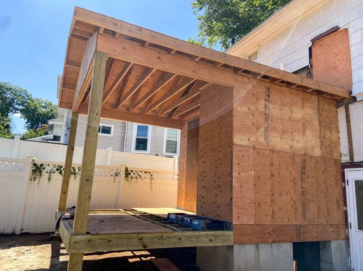 Wooden structure under construction, with plywood walls and a roof supported by wooden beams, partially built on a concrete foundation in a backyard with a white fence and neighboring houses in the background.