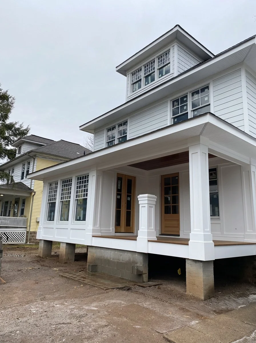 A new, white, multi-story house under construction with a porch and large windows, elevated on concrete and wooden supports, with a cloudy sky overhead.