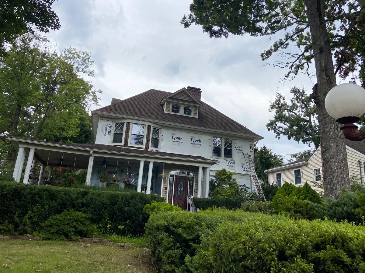 A two-story house under construction, with windows installed and Tyvek house wrap on the upper part, surrounded by green bushes and large trees, and a ladder leaning against the house.