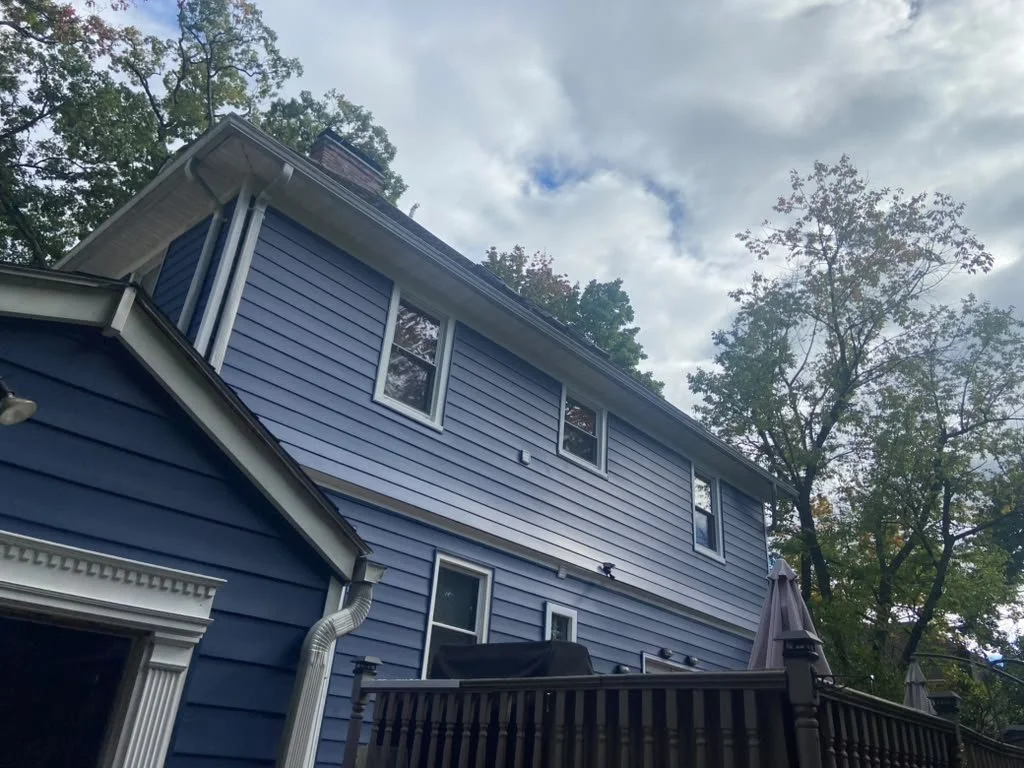 Back of a two-story blue house with a deck and trees overhead, partly cloudy sky.