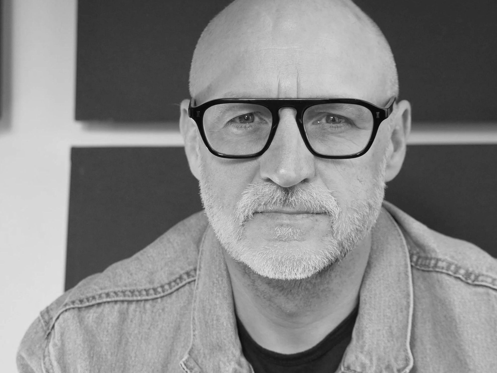 man with glasses, and a beard, wearing a denim jacket and a black shirt underneath.
