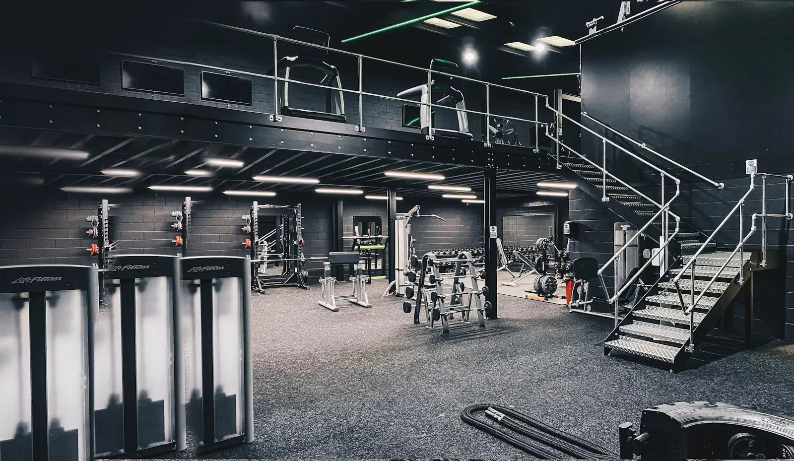 A modern gym with black walls and flooring, featuring various workout equipment including weight racks, benches, and cardio machines, with a staircase leading to a mezzanine level.