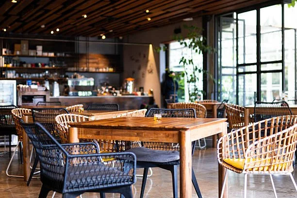 Interior of a modern coffee shop with wooden tables and woven chairs, large windows, and a counter in the background.