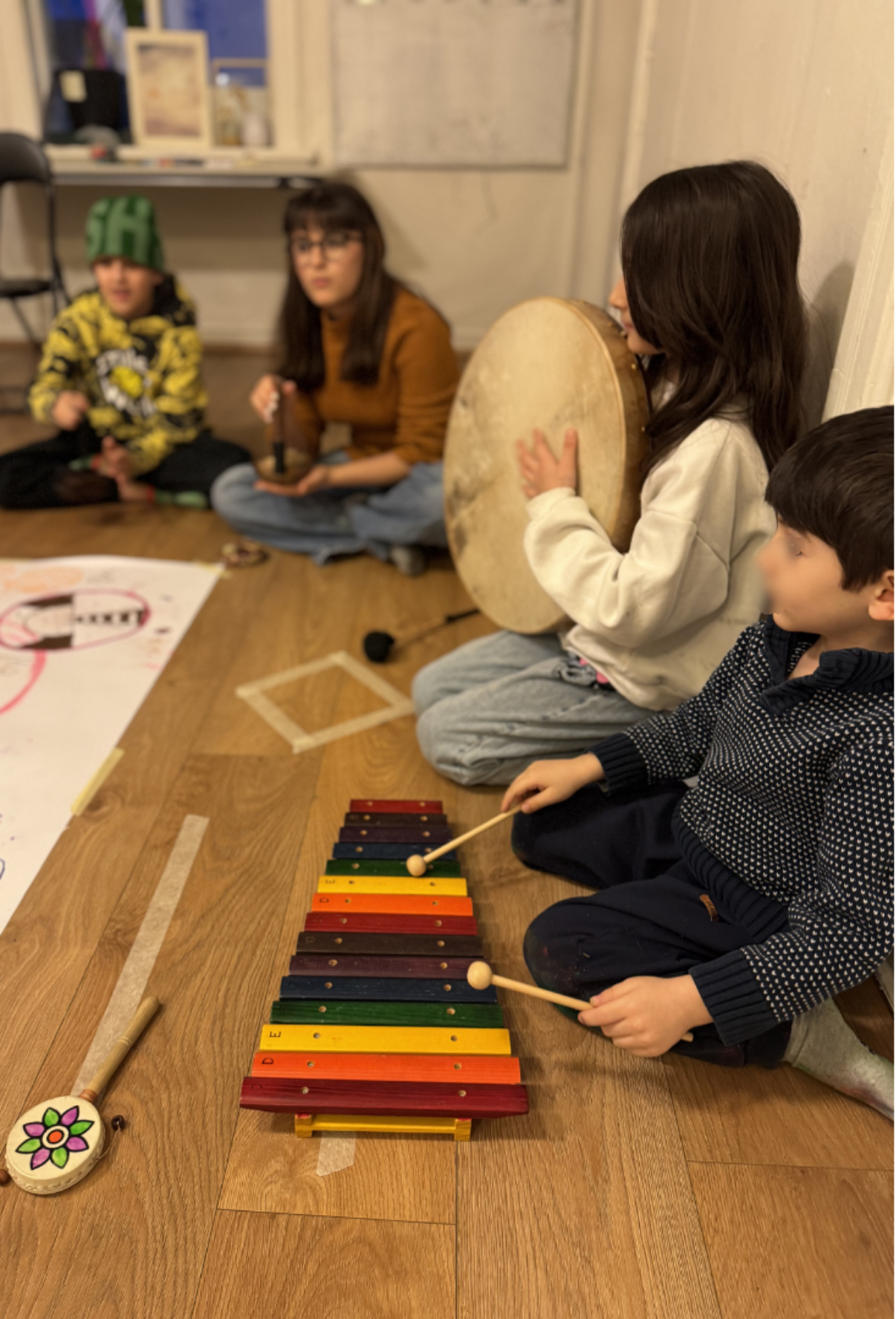 Four children sitting on the floor in a room, playing musical instruments, with a colorful xylophone on the floor in front of them.