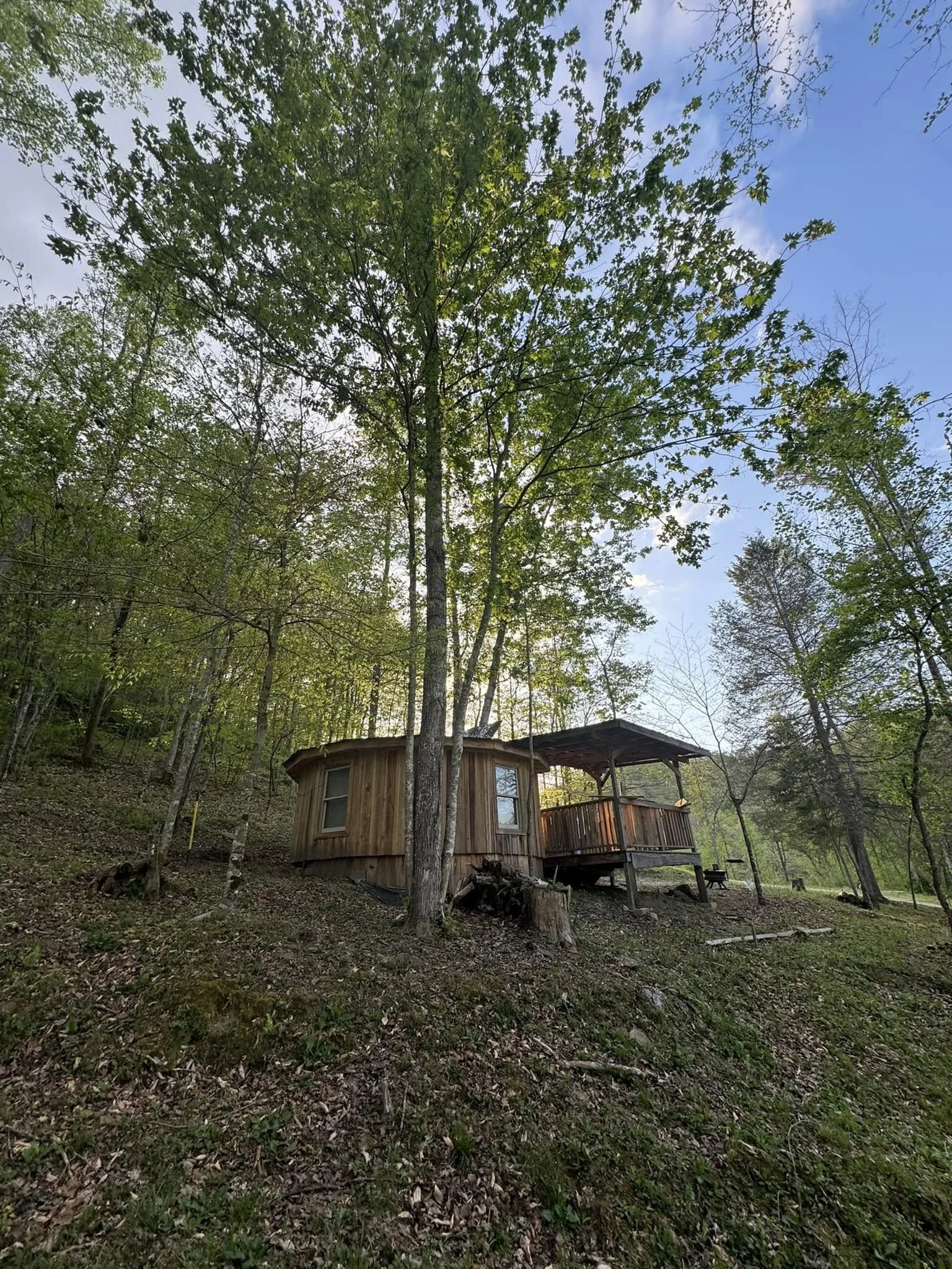 A small, round wooden cabin with a covered porch, surrounded by tall green trees and a forested area, under a partly cloudy sky.