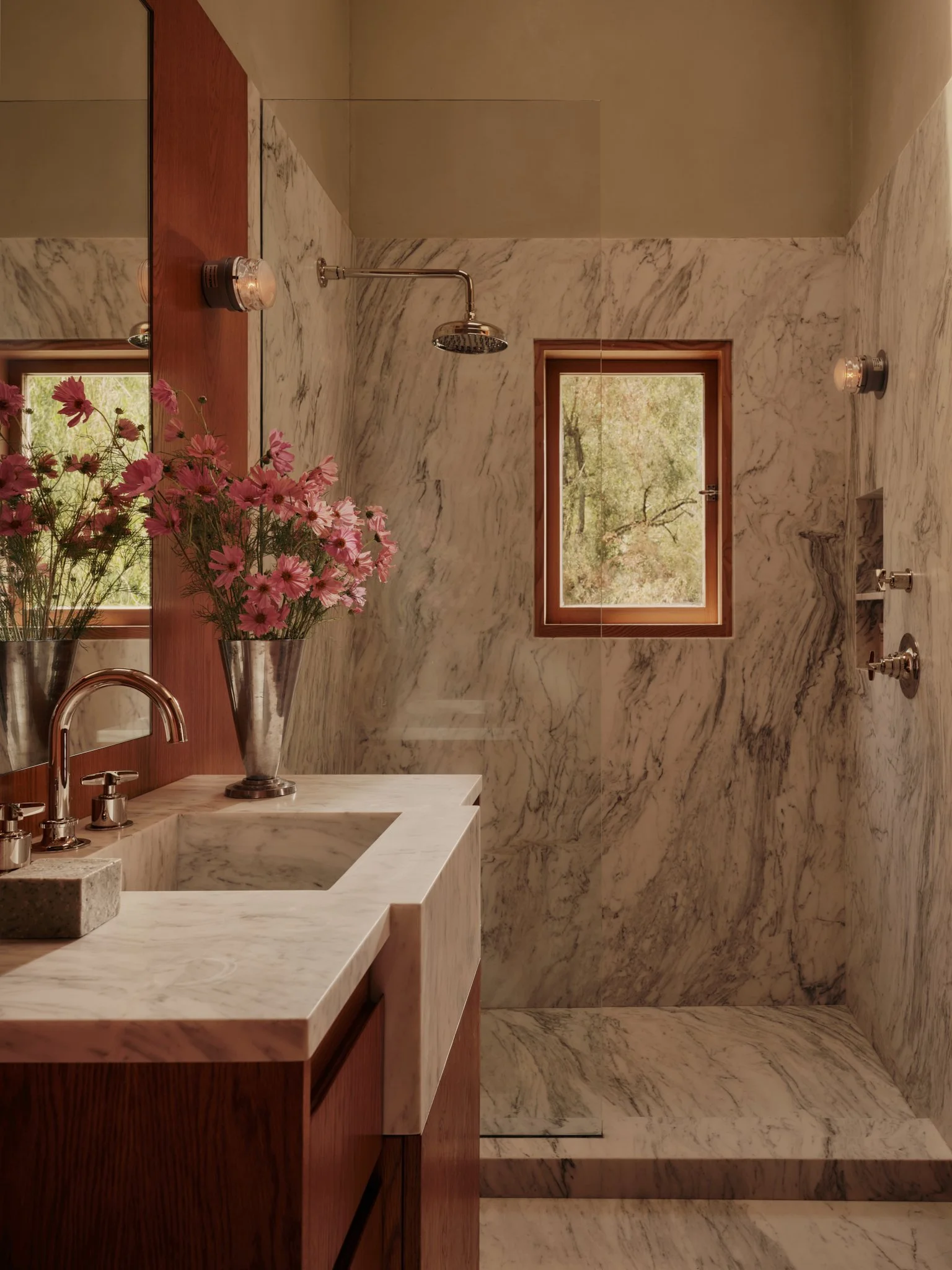 A bathroom with marble walls and floors, a wooden vanity with a white marble countertop, a pink flower bouquet in a silver vase, a window showing trees outside, a showerhead, and a mirror.