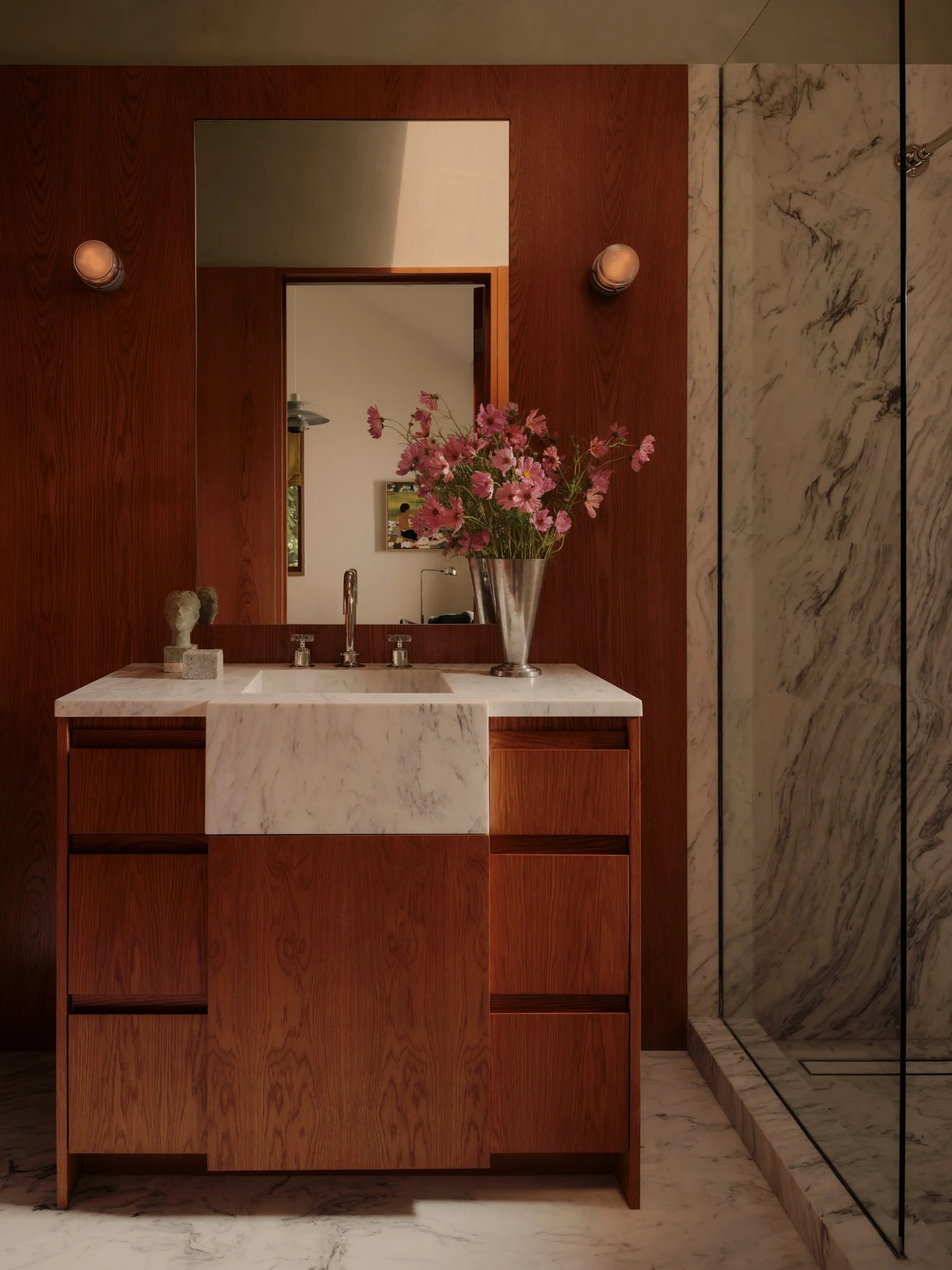 A bathroom vanity with a marble countertop, a large mirror, and a wooden cabinet. A vase with pink flowers sits on the counter, and wall-mounted lights are on either side of the mirror. The background includes a marble shower wall.