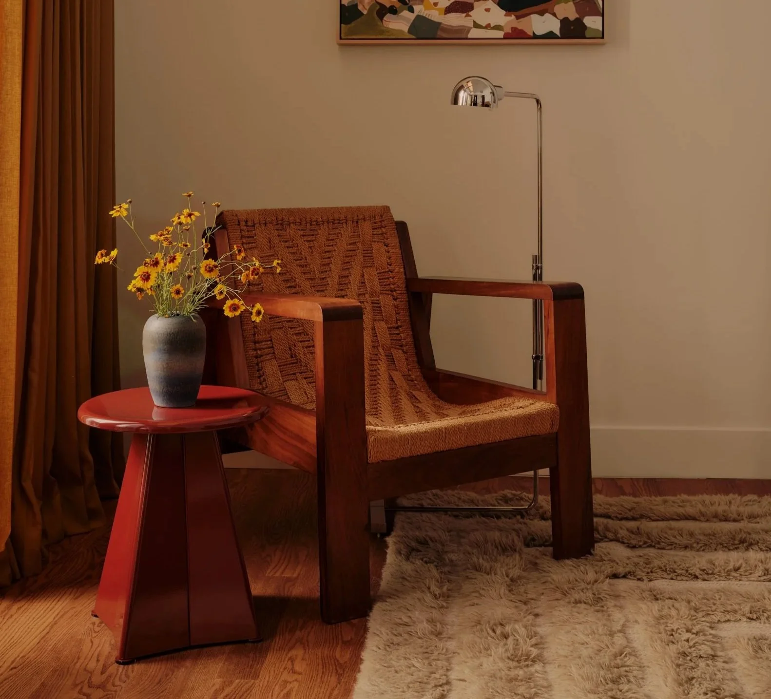 A cozy room corner featuring a wooden armchair with a woven cushion, a small round side table with a vase of yellow flowers, a floor lamp with a metallic shade, a wooden floor, a cream-colored wall, a multicolored abstract artwork, and a plush beige shaggy rug.