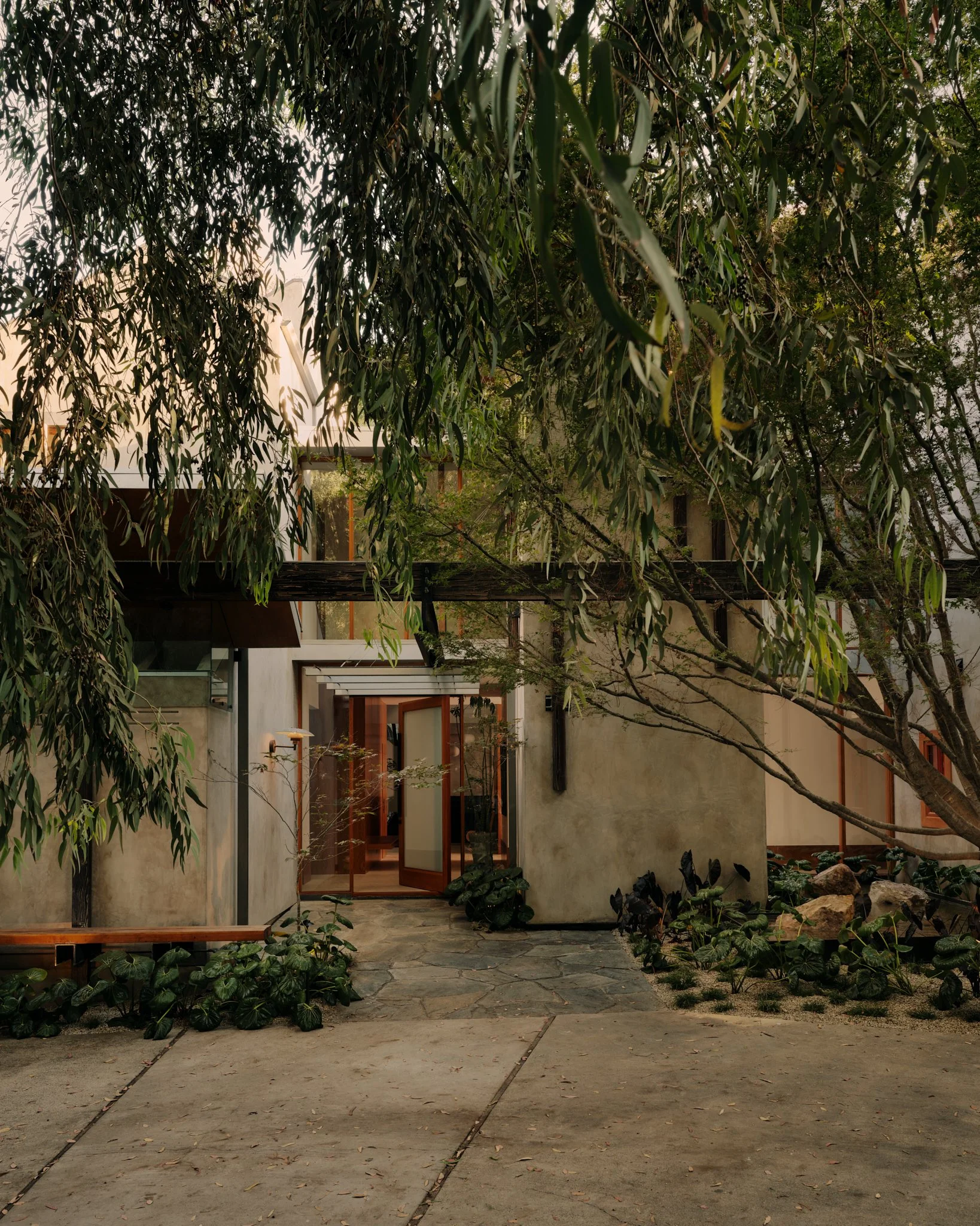 A modern house with large glass doors, surrounded by lush green trees and plants in a garden with stone pathway.
