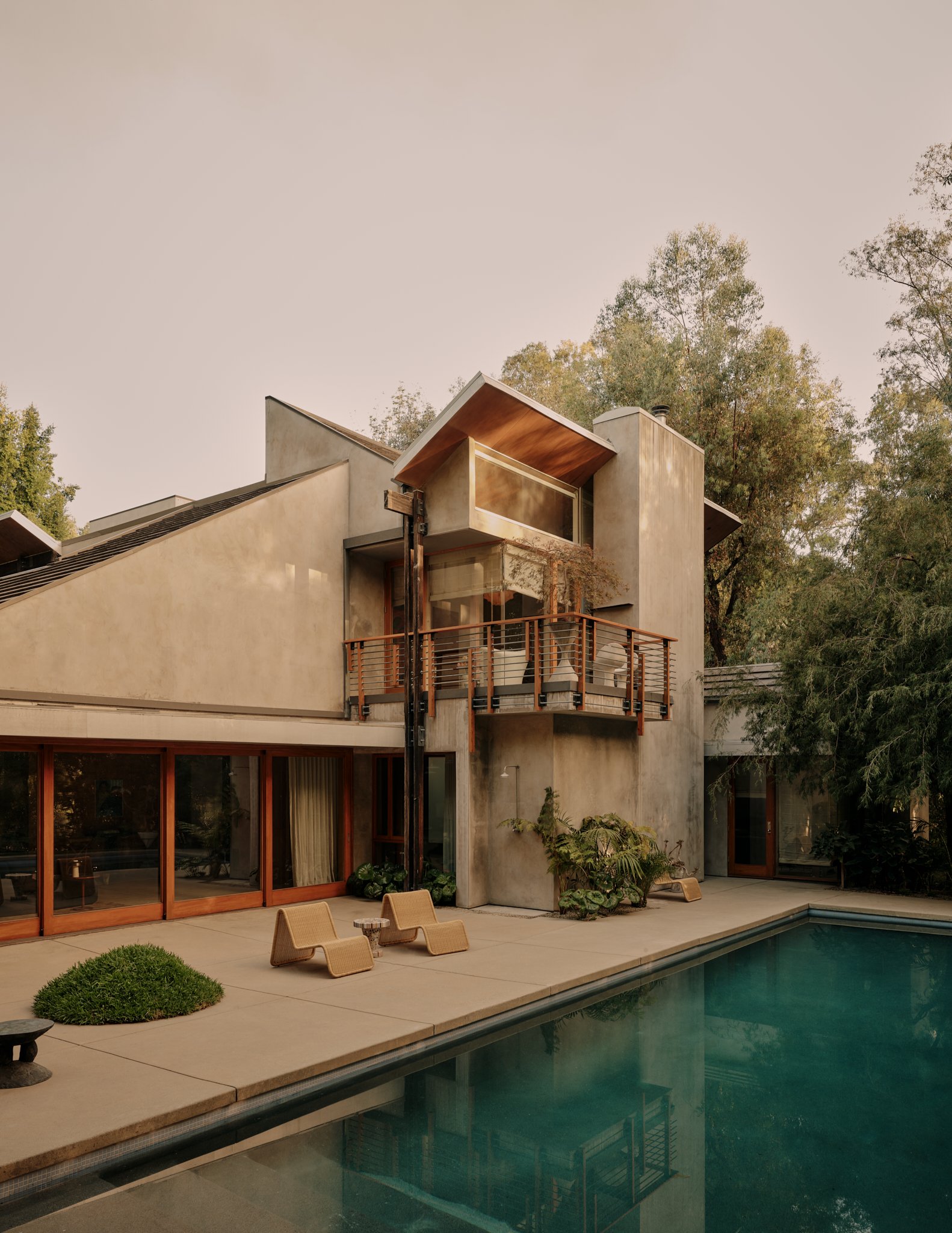 Modern house with large glass sliding doors, outdoor patio furniture, a swimming pool, and trees in the background during daylight.