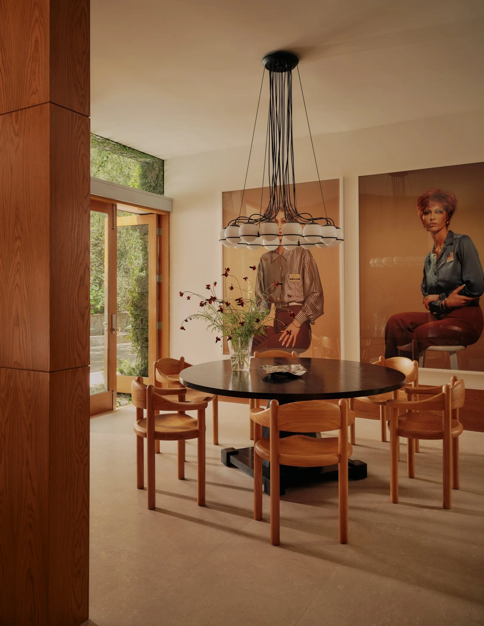 A room with a round black table, wooden chairs, a vase with flowers, large posters of women, and a chandelier hanging from the ceiling.
