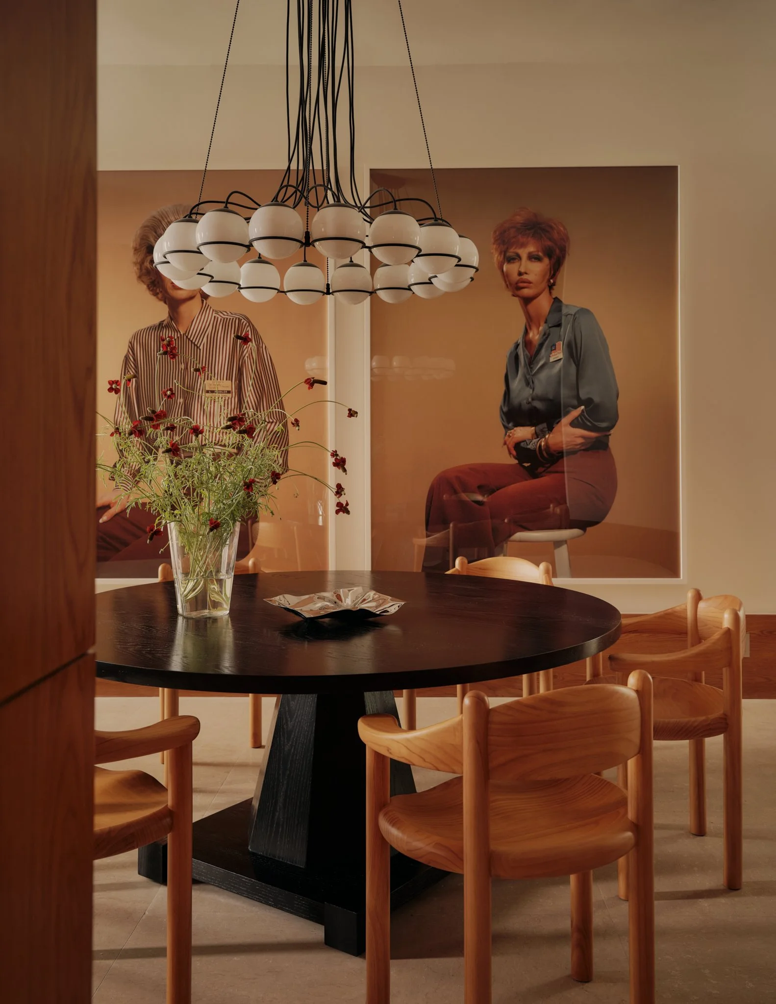 A round black dining table with light wood chairs surrounding it, a glass vase with red flowers on the table, and a large piece of artwork featuring two women, one with short reddish hair in a blue shirt and the other with hair obscured behind a chandelier, hanging on the beige wall in the background.
