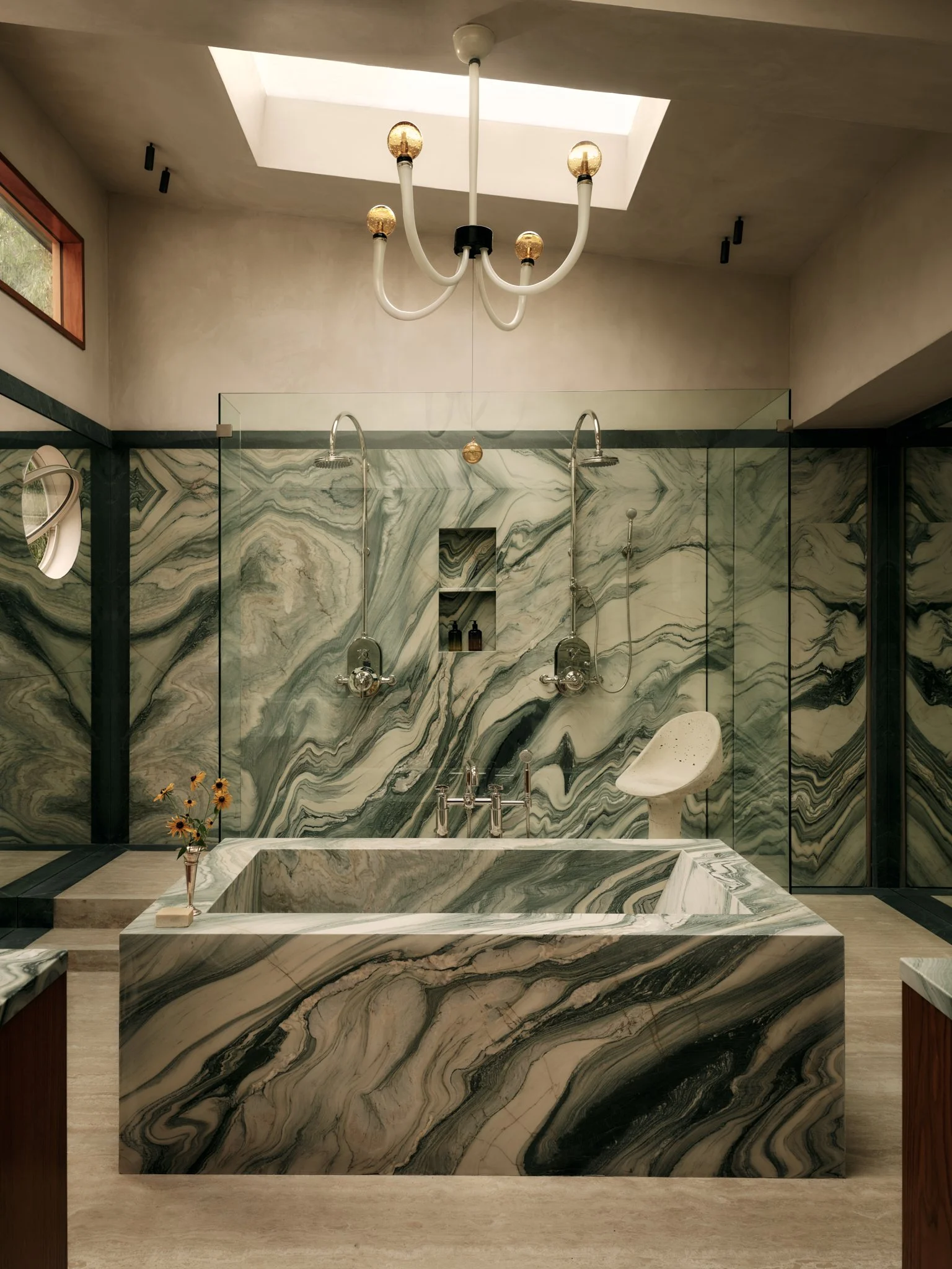 Bathroom with a large marble bathtub, a marble wall with shower heads, a small white chair, a vase with flowers, and a window with black trim. A modern chandelier hangs from the ceiling.