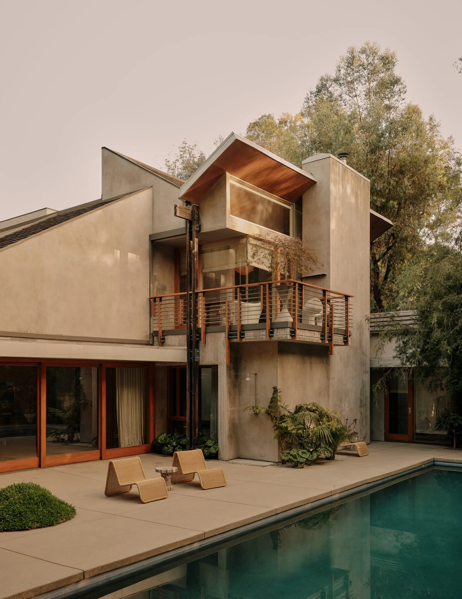 A modern house with large glass doors opening to a backyard pool. There are wooden lounge chairs and patio plants near the pool.