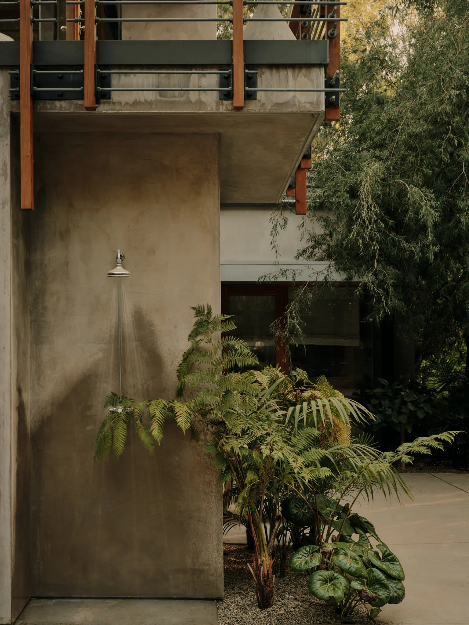 An outdoor shower with a metal nozzle on a concrete wall, surrounded by lush green plants and trees in a garden setting.