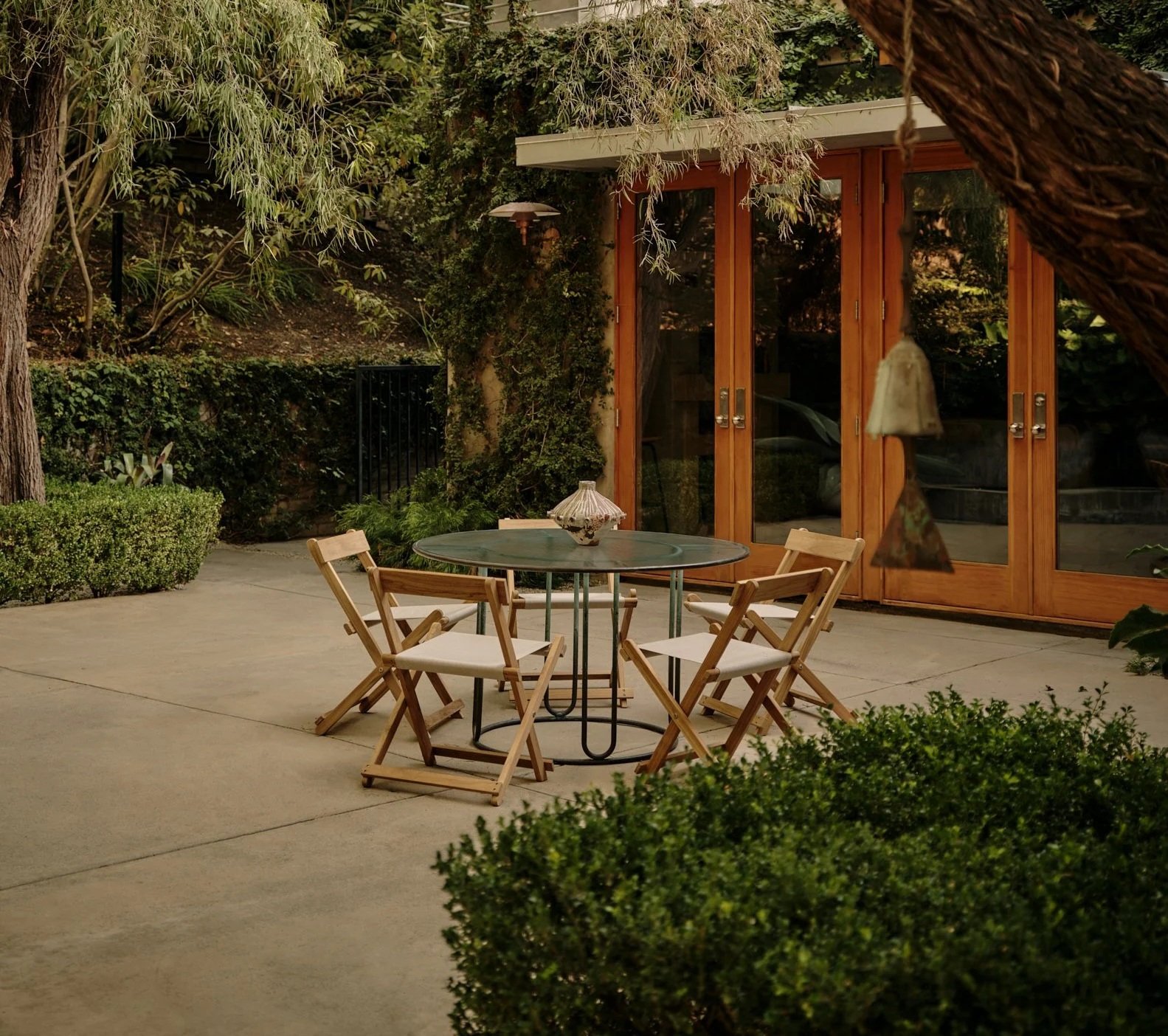 An outdoor patio with a round glass table, four wooden chairs with white cushions, a decorative lamp on the table, and a background of lush greenery and wooden-framed glass doors.