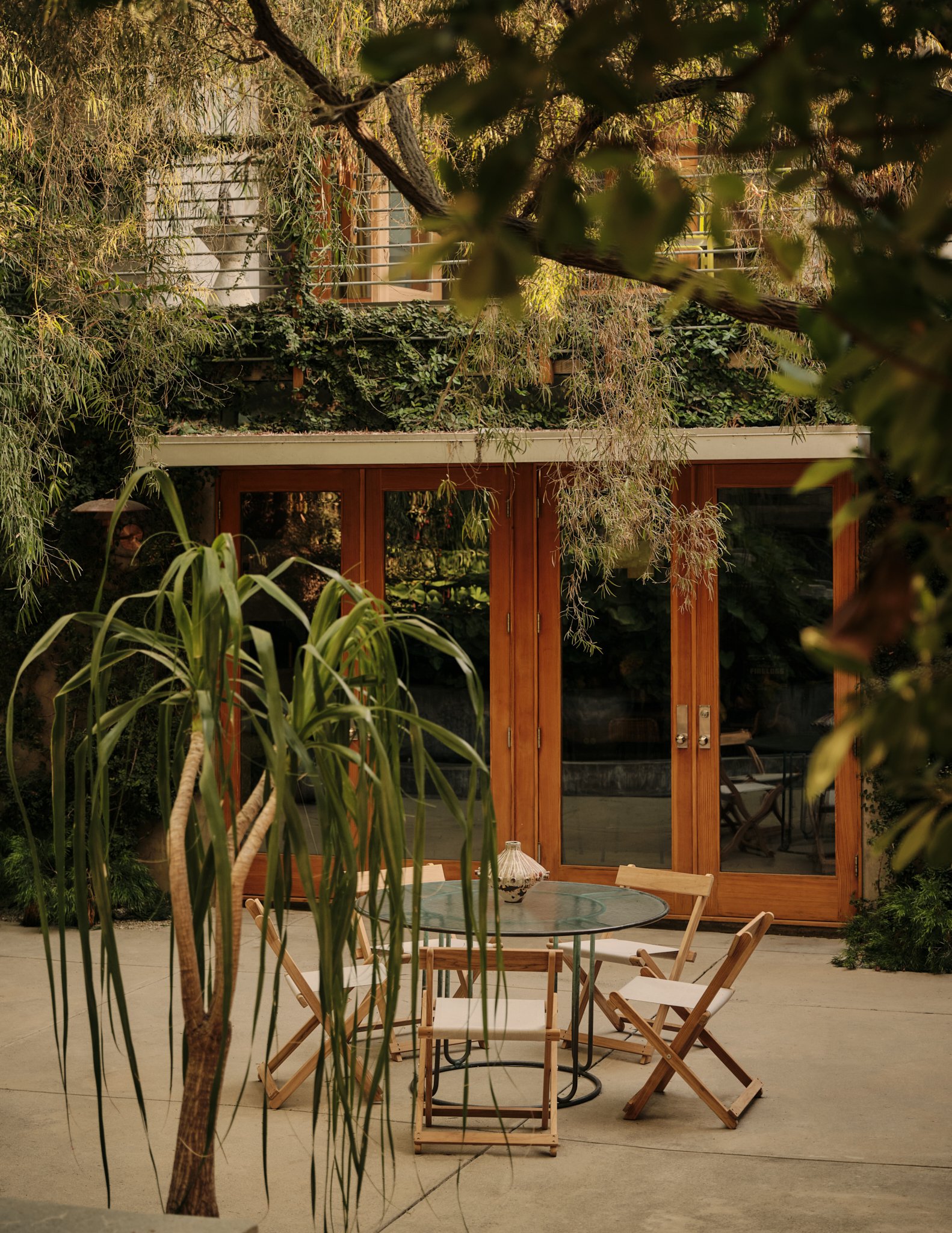 An outdoor patio area with wooden and metal furniture, surrounded by plants and trees, with a wooden house in the background.