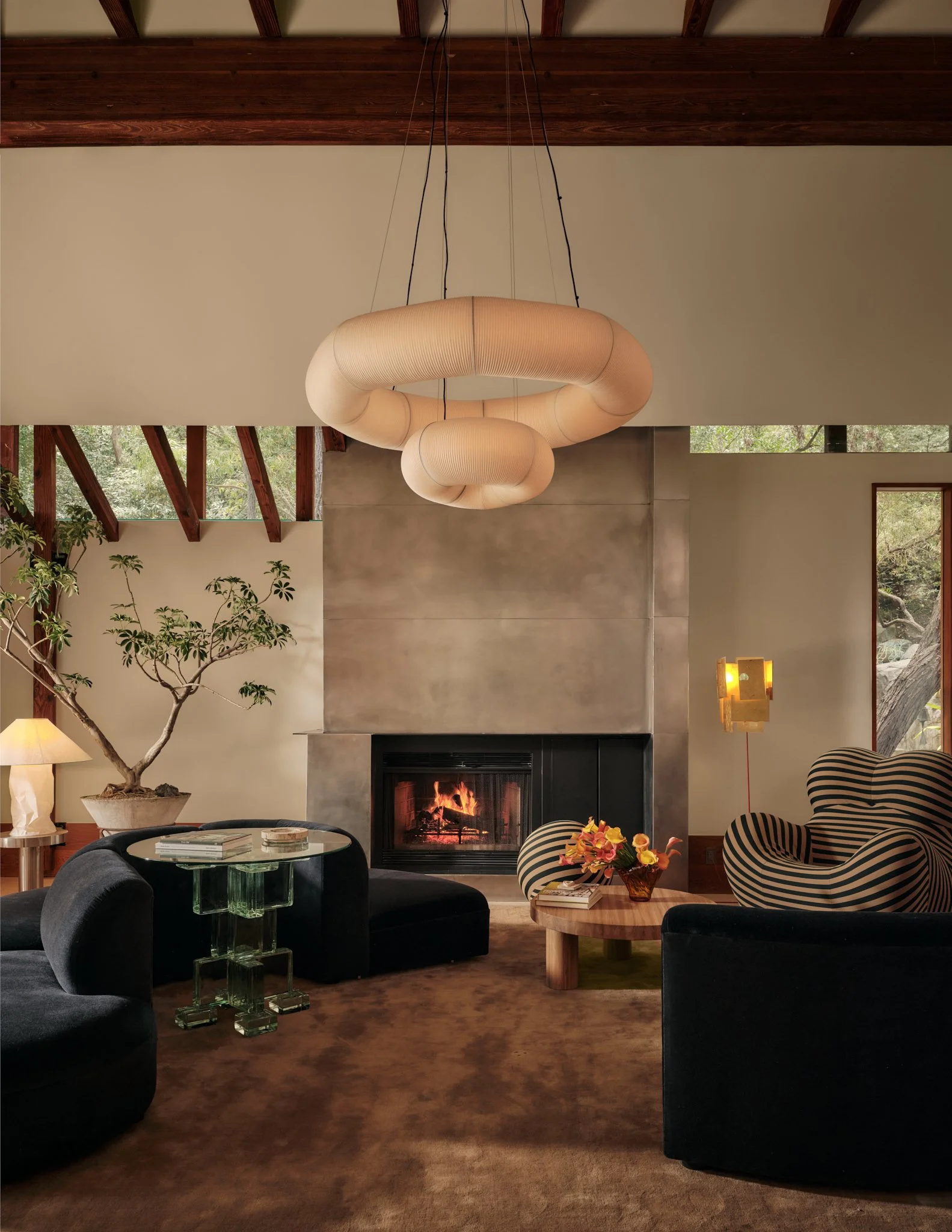 Living room with black armchairs, a glass side table, a fireplace, a potted plant, and modern lighting fixtures, featuring wood beams on the ceiling and large windows showing outdoor greenery.
