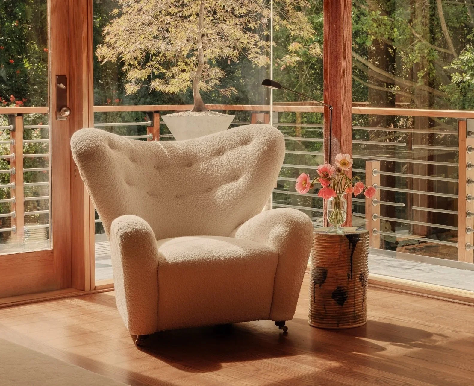 Cozy living space with a cream-colored armchair, a side table with a vase of pink flowers, and a large window showing trees outside.