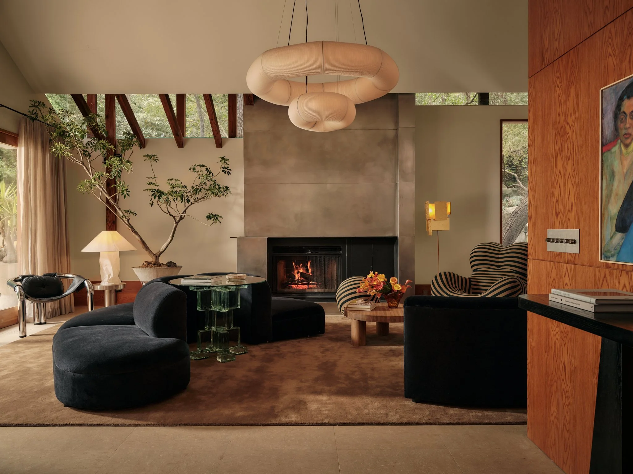 A modern living room with a fireplace, a large bonsai tree, black curved sofas, glass coffee tables, artwork, a lamp, and large windows showing trees outside.