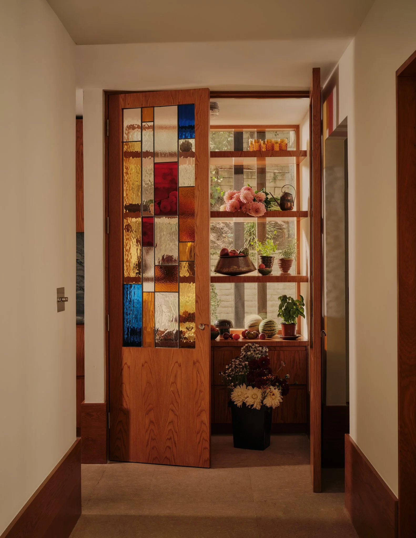 A wooden partition with stained glass panels separates a hallway from a room with a window. The room features shelves with decorative items and flowers, including a large bouquet of pink peonies and red, white, and green plants in pots.