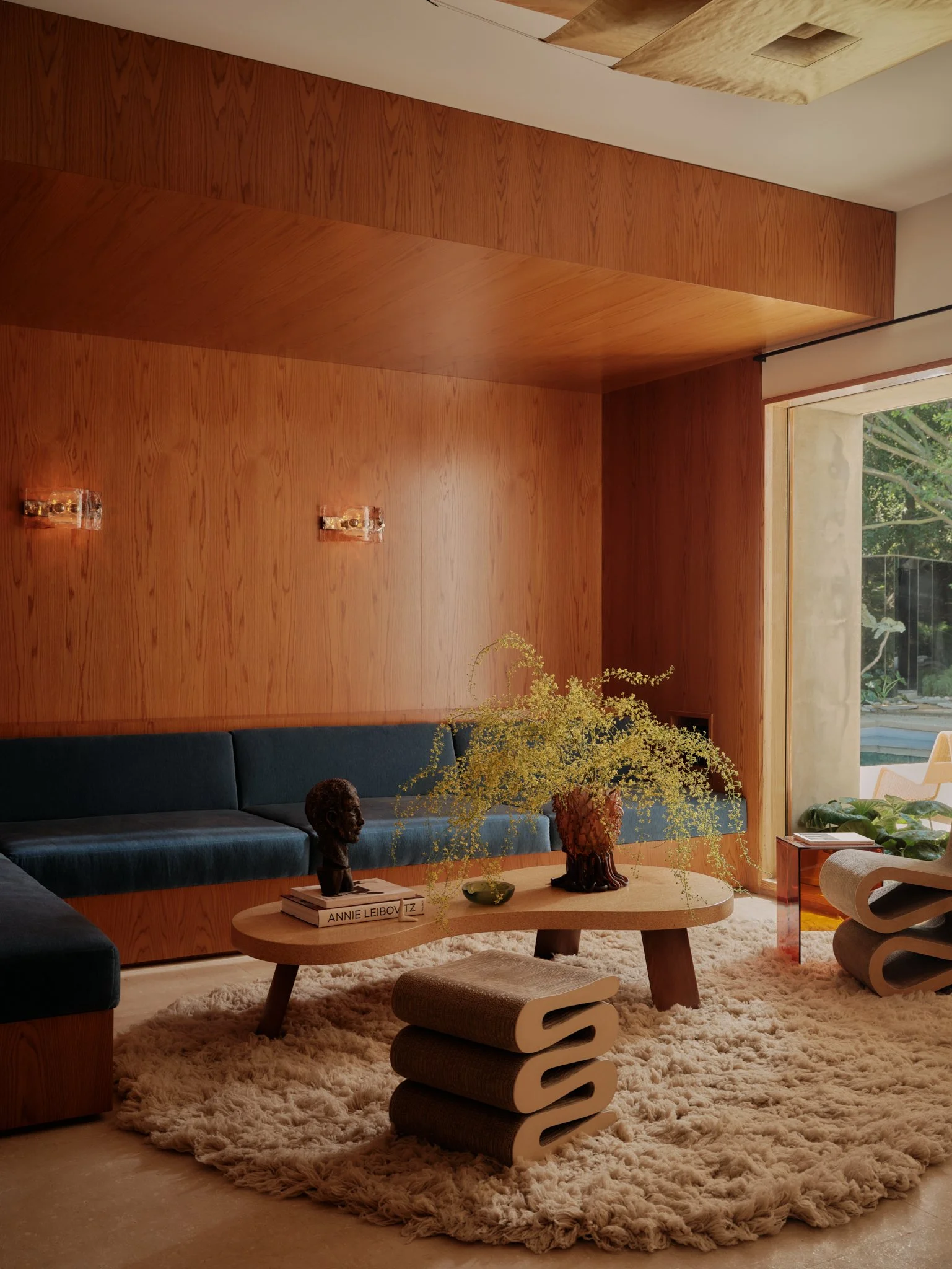 A modern living room with a wooden wall, a blue sofa, a round wooden coffee table with decorative items, a plush shaggy beige rug, and a large window with a view of greenery outside.