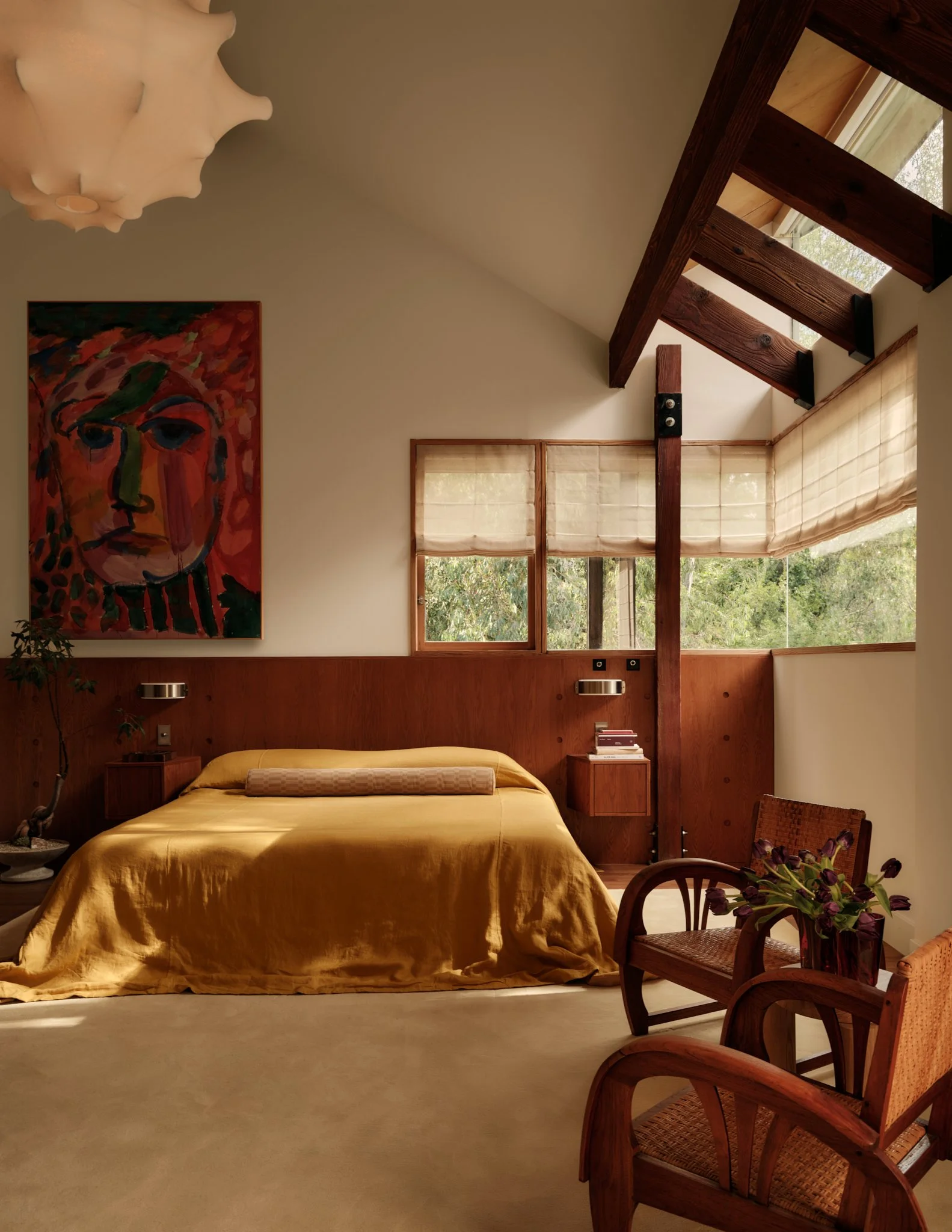 A bedroom with a bed covered in a yellow silky bedspread, a wooden headboard, and a modern ceiling light. There are two windows with beige blinds, a colorful abstract portrait painting on the wall, and a wooden chair with a woven seat holding a vase of purple flowers. Wooden beams are visible on the ceiling, and the room has a cozy, mid-century modern style.