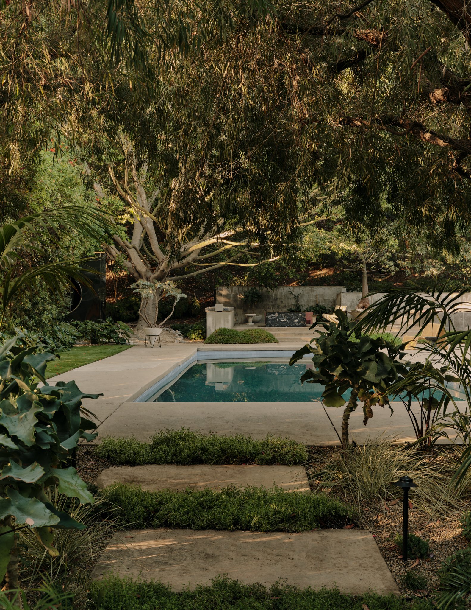 A backyard with a swimming pool, surrounded by lush green plants and trees, with an outdoor seating area and a low fence.