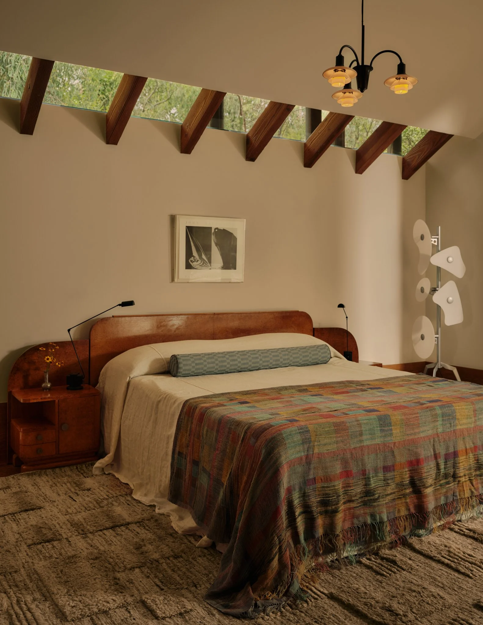 A bedroom with a bed that has a wooden headboard, beige bedding, and a multiple-color plaid throw blanket. There is a bedside table with a small vase and a black lamp. Above the bed, there are two framed black and white pictures on the wall. The room features a high ceiling with angled wooden beams spaced in a row, with a strip of windows between the beams allowing natural light. A vintage-style chandelier with five lights hangs from the ceiling. In the corner of the room, there is a modern floor lamp with white, flower-shaped shades.